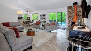 A spacious living room with a grey sofa, a coffee table, and a fireplace. There is a dining area with chairs and a table, and a kitchen in the background. - Alejo Olive Orchard- Beautiful 3 Bed Home with Pool/Spa  - 5 minute drive to downtown Palm Springs. (Palm Springs)
