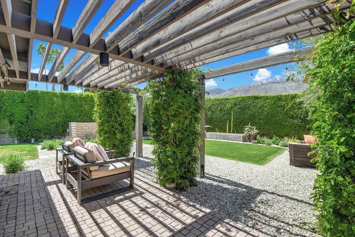 A covered patio with a wooden pergola, a seating area with cushions, and a well-maintained garden with hedges and a lawn.