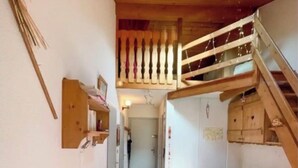 En smal hall med en trätrappa som leder upp till en övre våning. En röd, mönstrad soffa står i hallen, och ett litet bord med stolar är placerat på den övre våningen.