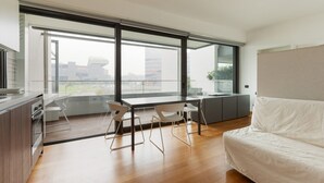 A modern room with a large window, a dining table with chairs, a sofa, and a kitchen area. - GuestHost - Cozy Studio in Portello District (Milano)