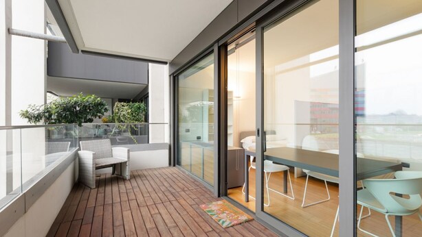 A balcony with a wooden floor, a wicker chair, a small table, and a glass door leading to an indoor area with a table and chairs. - GuestHost - Cozy Studio in Portello District (Milano)