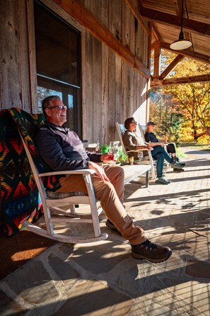 A wooden porch with two rocking chairs and a table, overlooking a garden with autumn foliage.
