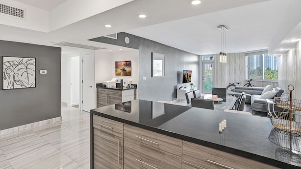A modern kitchen with a large island, a dining area with a sofa and chairs, and a view of the city through large windows. - Sunny Isles Resort by the beach w/ Pool, Games & Gym (Sunny Isles Beach)