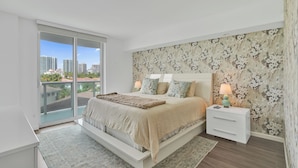 A bedroom with a large bed, a nightstand, a lamp, a view of the city through a balcony. - Sunny Isles Resort by the beach w/ Pool, Games & Gym (Sunny Isles Beach)