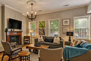 A living room with a fireplace, a television, and a chandelier. - Park Place, Unit A - 2BR near Forsyth w/Balcony, Courtyard, Easy Street Parking (Savannah)