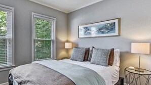 Une chambre à coucher avec un lit, deux lampes de chevet, un tableau encadré au mur et une fenêtre avec des stores.