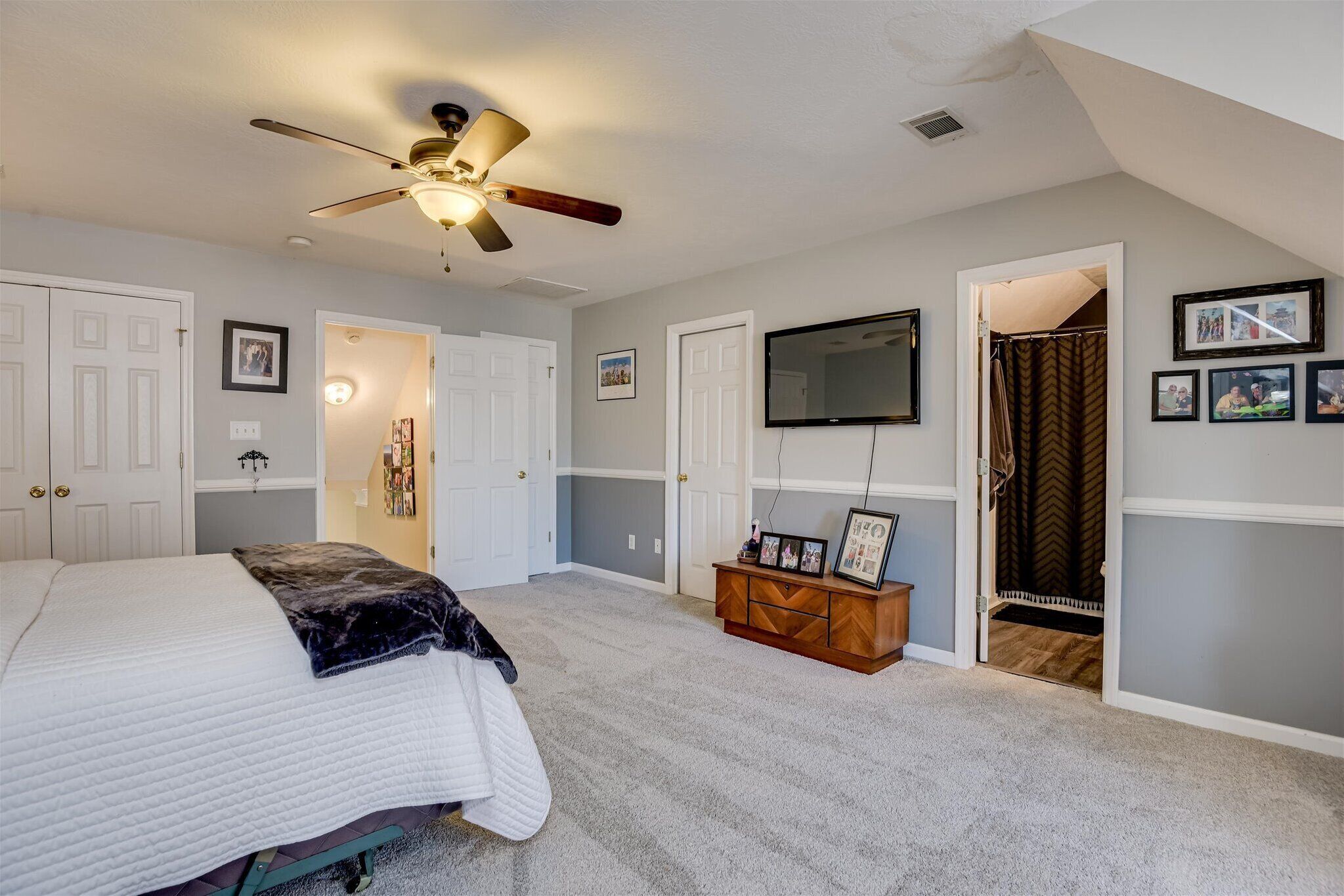 A bedroom with a bed, a ceiling fan, a TV mounted on the wall, and a wooden dresser.