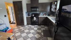 Une cuisine avec des meubles de rangement en bois foncé, un réfrigérateur noir, un four à micro-ondes et un sol carrelé agrémenté d’un tapis à motifs.