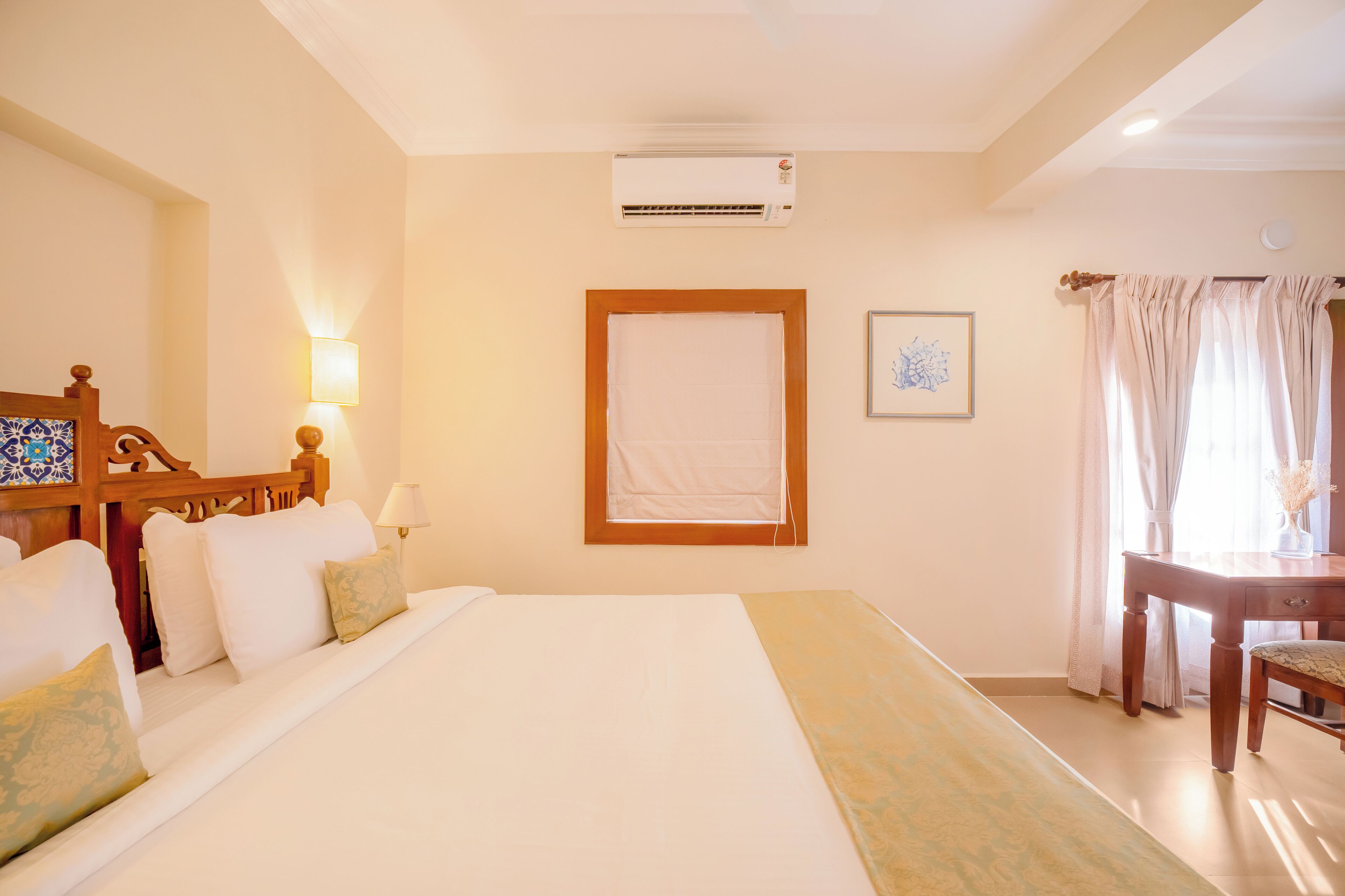 Una habitación de hotel con una cama grande, cabecera de madera, un escritorio con silla, una ventana con cortinas y un equipo de aire acondicionado en la pared.