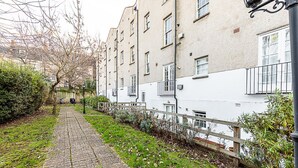 Ein schmaler Pflasterweg, gesäumt von Gebäuden mit weißen und beigen Fassaden, einem Holzzaun und einem Laternenpfahl.