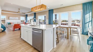 Uma cozinha moderna com lava-louças, uma sala de jantar com mesa e cadeiras de madeira, e uma sala de estar com sofá e uma cadeira vermelha.
