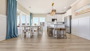 Uma cozinha ampla com armários brancos, uma área de jantar com mesa e cadeiras, e uma janela grande com cortinas.