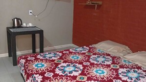 Een slaapkamer met een bed, een klein tafeltje met een waterkoker, een ventilator en een aan de muur bevestigde televisie.