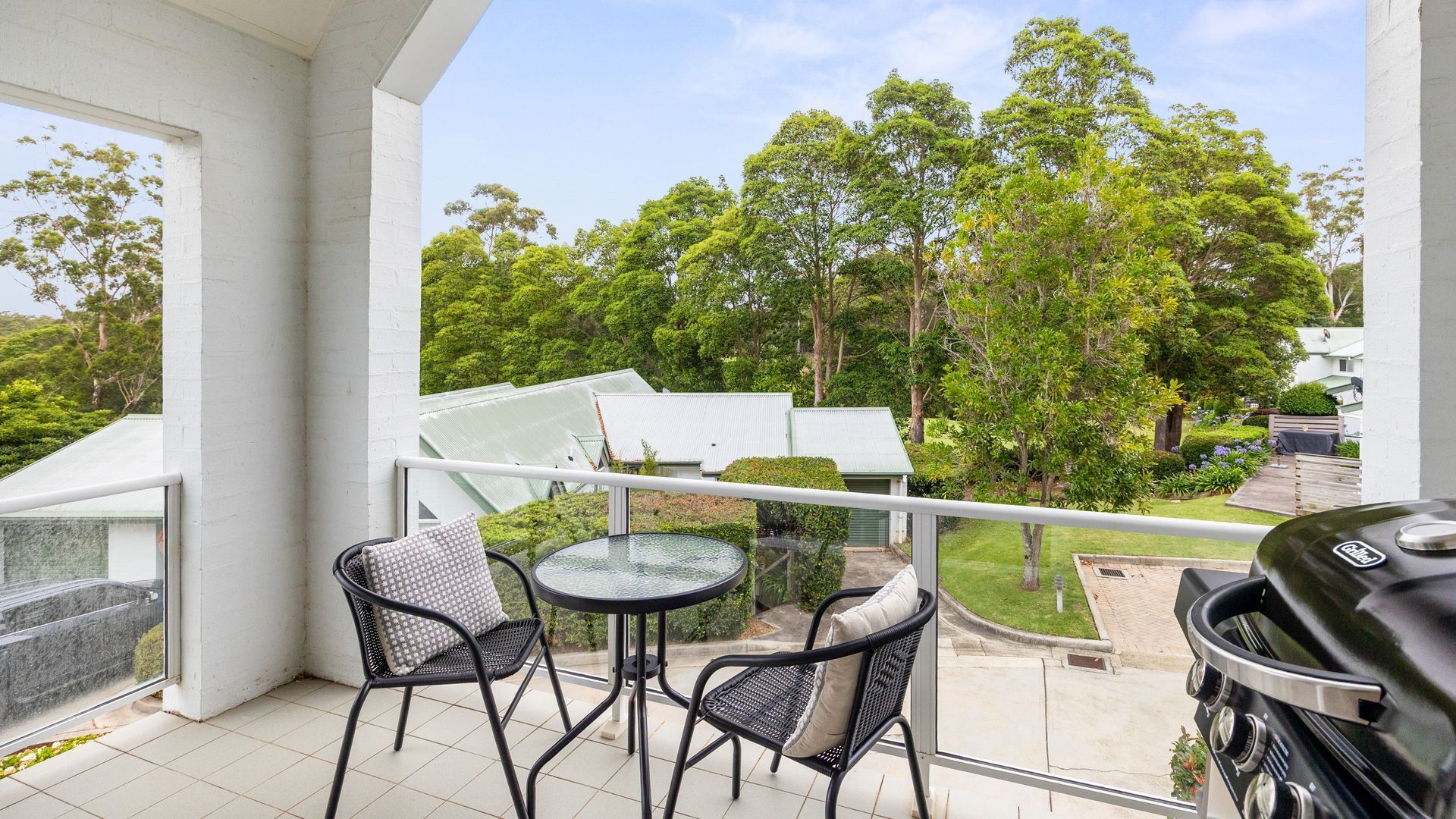 A balcony with two chairs and a table, overlooking a garden with trees and a barbecue grill.
