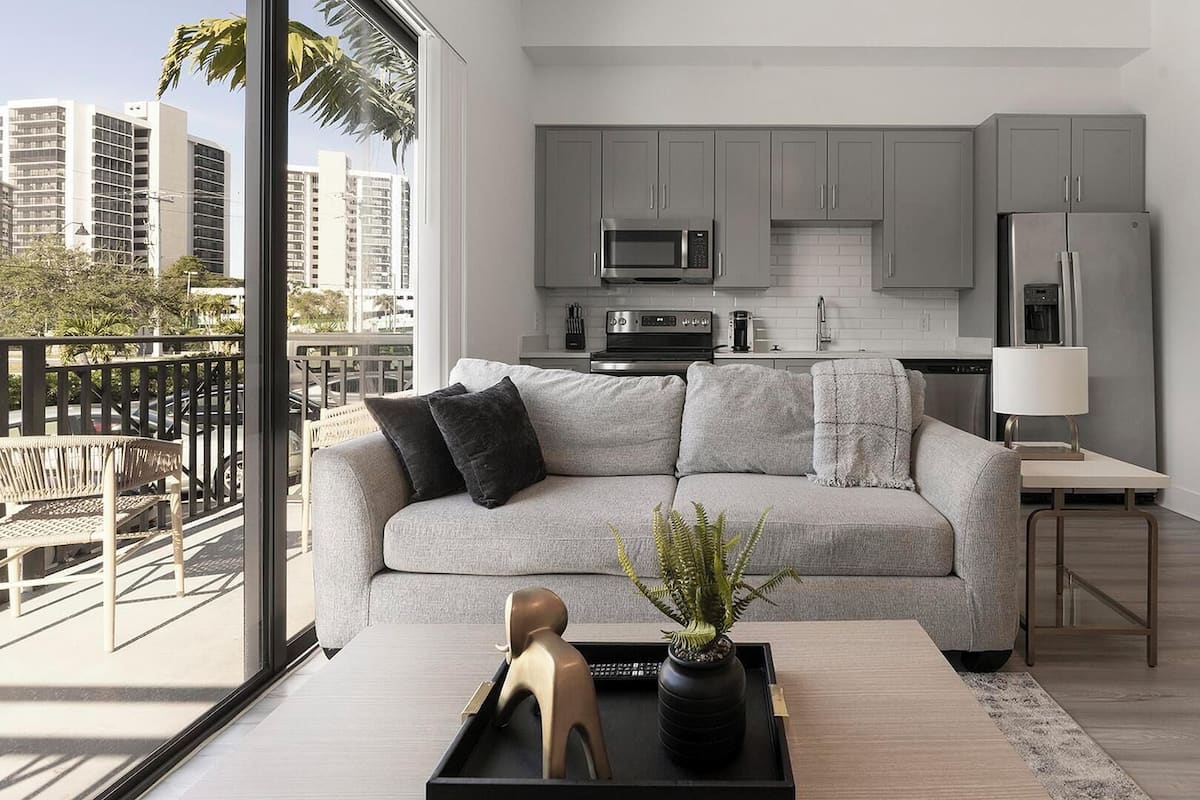 A modern apartment with a grey sofa, a dining area, a kitchen with grey cabinets, and a view of palm trees and buildings.