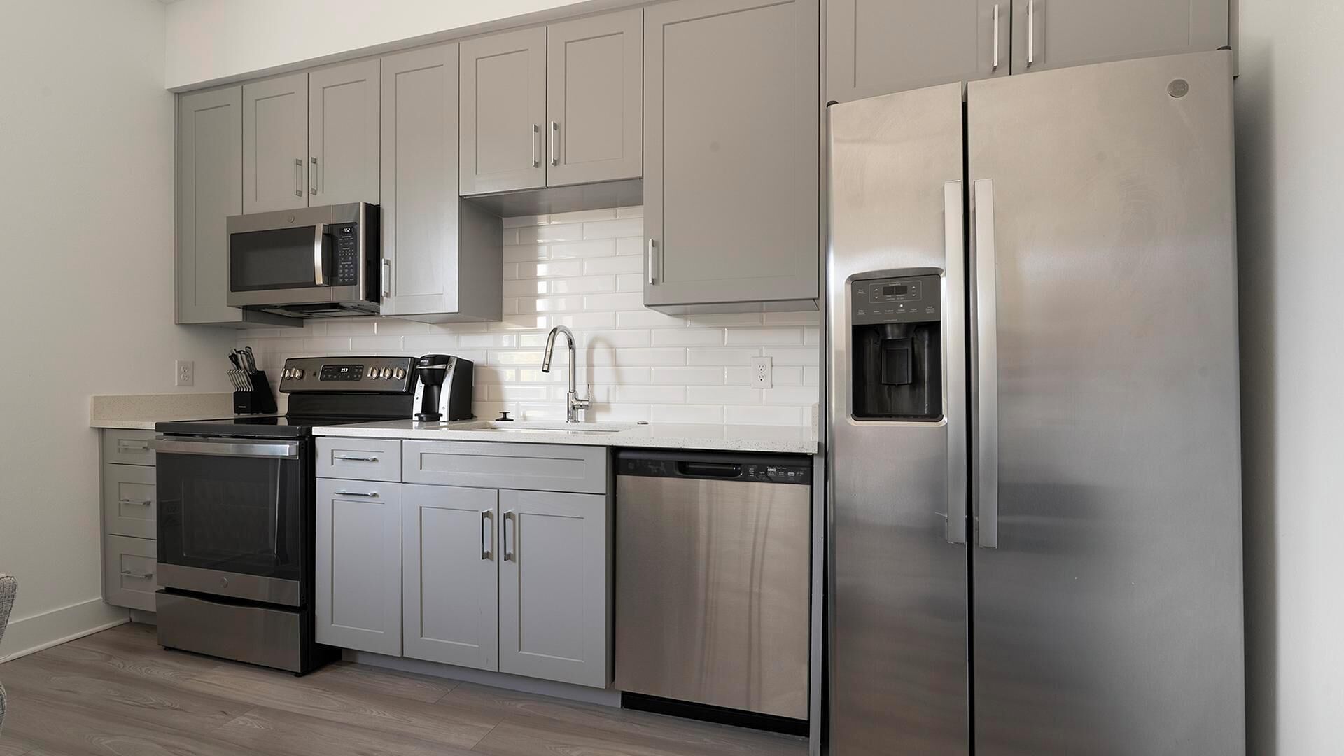 A modern kitchen with stainless steel appliances, grey cabinets, and a white tiled backsplash.