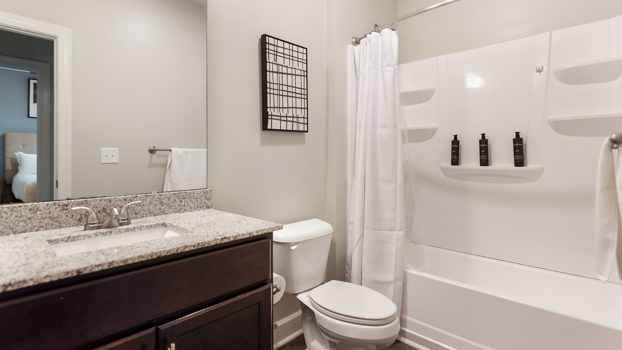 A bathroom with a granite countertop sink, a toilet, and a bathtub with a shower curtain. There are three bottles on the shelf above the bathtub.