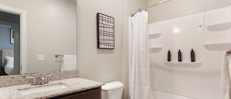A bathroom with a granite countertop sink, a toilet, and a bathtub with a shower curtain. There are three bottles on the shelf above the bathtub.