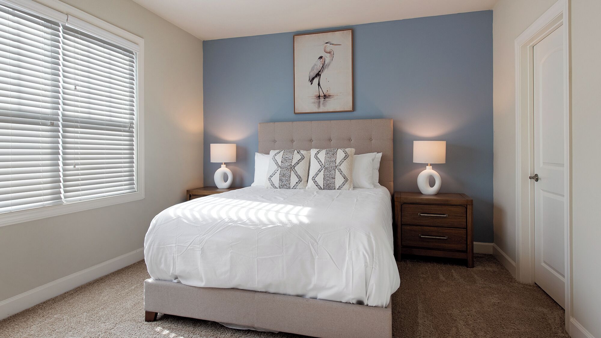 A bedroom with a bed, two bedside tables with lamps, a nightstand, a framed picture on the wall, and a window with blinds.