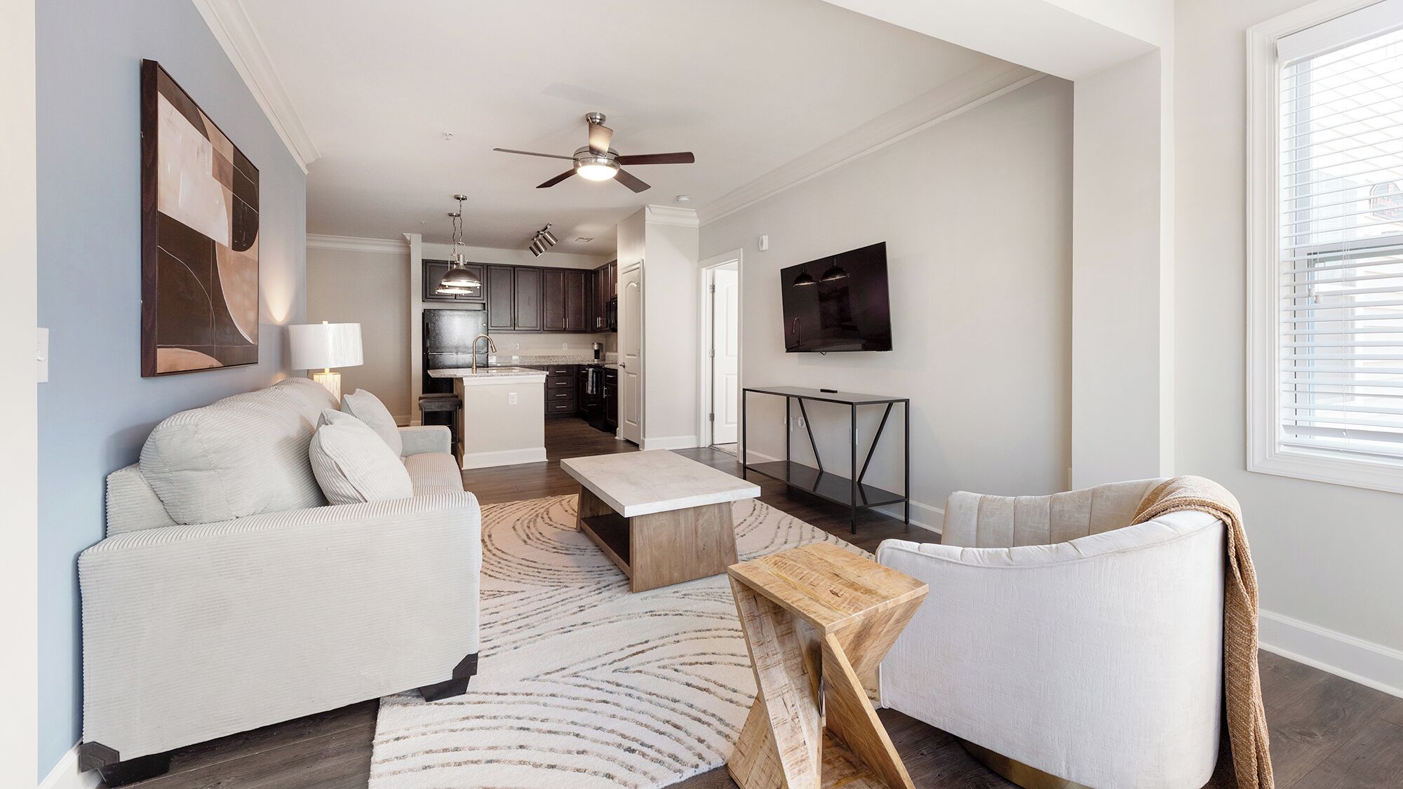 A modern living room with a sofa, coffee table, and TV, connected to a kitchen with dark cabinets and a ceiling fan.