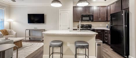 A modern kitchen with dark wood cabinets, a black refrigerator, a microwave, and a stainless steel sink. There are two bar stools at the island, and a flat-screen TV mounted on the wall.