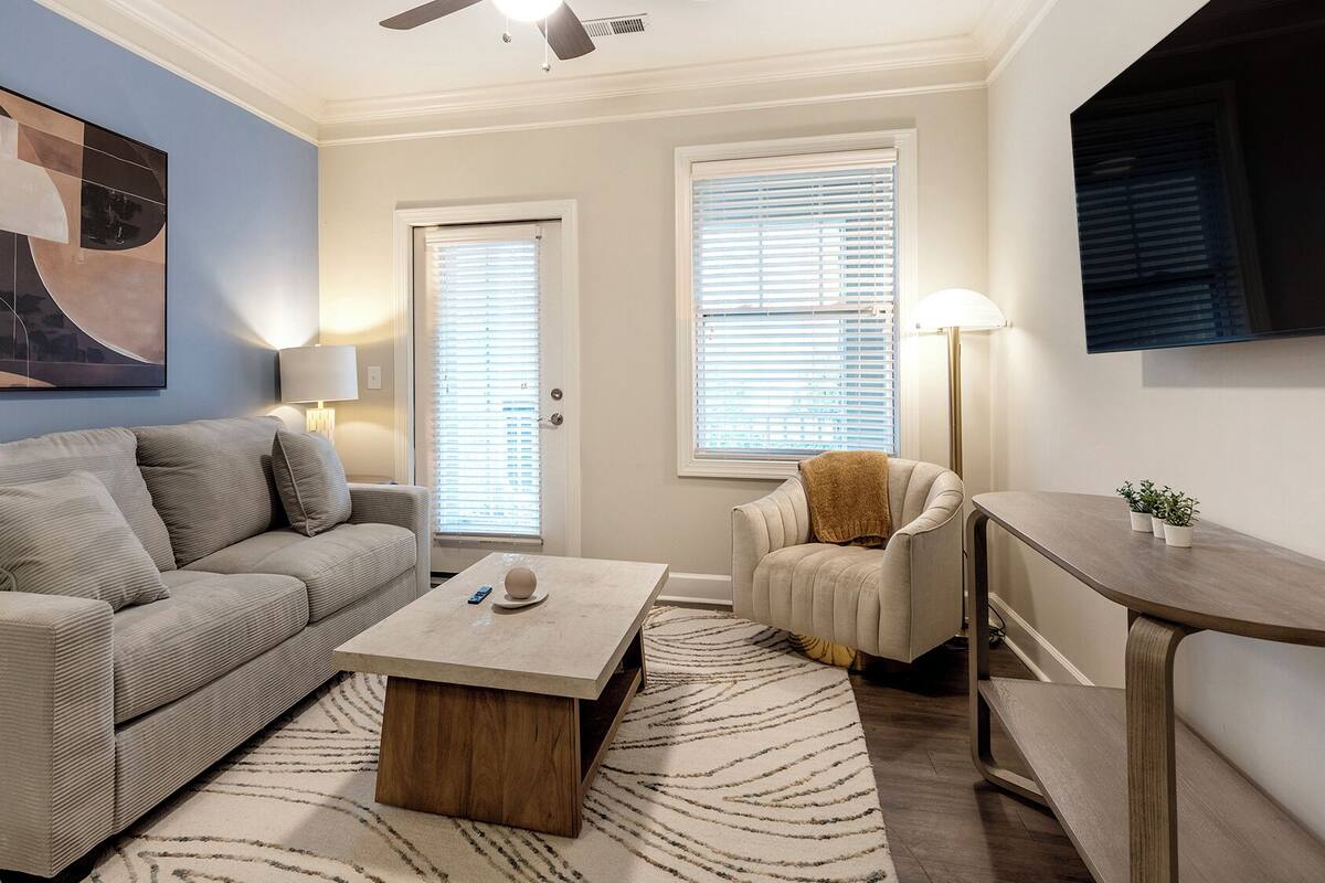 A living room with a sofa, coffee table, and armchair, a painting on the wall, and a television mounted above a wooden table.