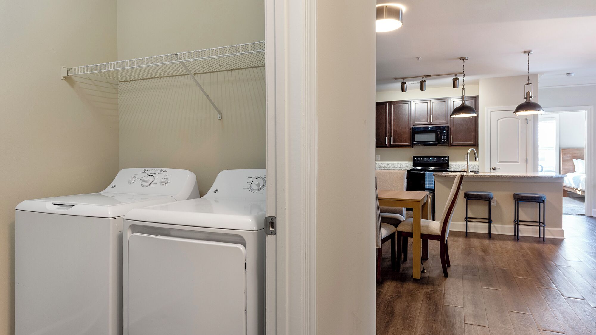 A laundry area with two white washing machines, a metal drying rack, and a kitchen with wooden cabinets, a microwave, and a dining area with stools.