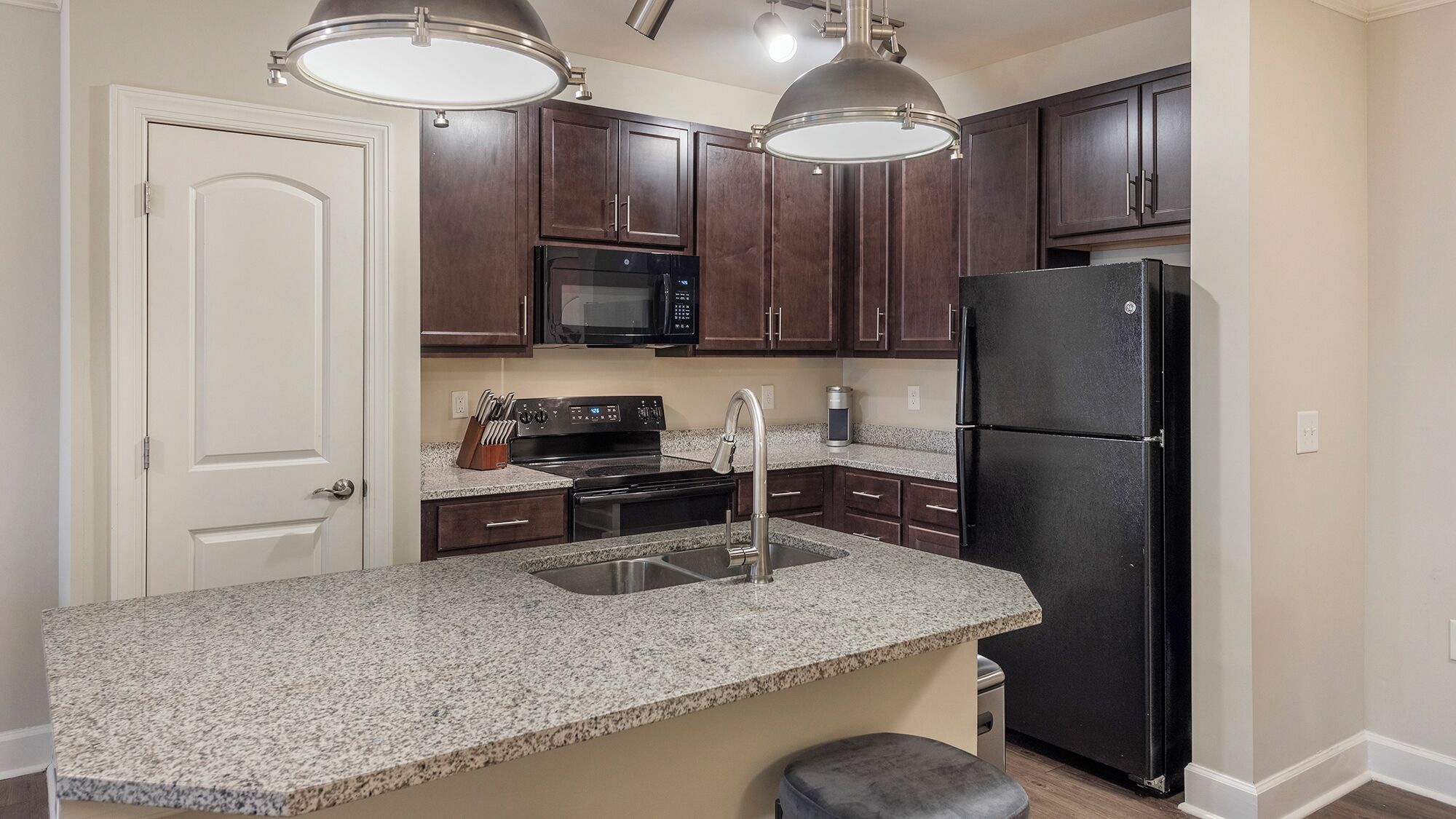 A kitchen with dark wooden cabinets, a black refrigerator, a microwave, and a stainless steel sink.