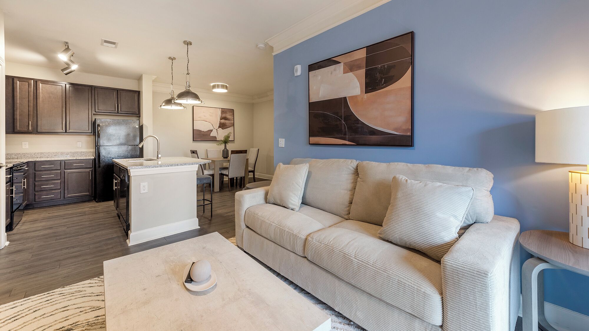 A modern kitchen with dark cabinets and a central island, a living area with a beige sofa and a coffee table, and a dining area with a table and chairs.