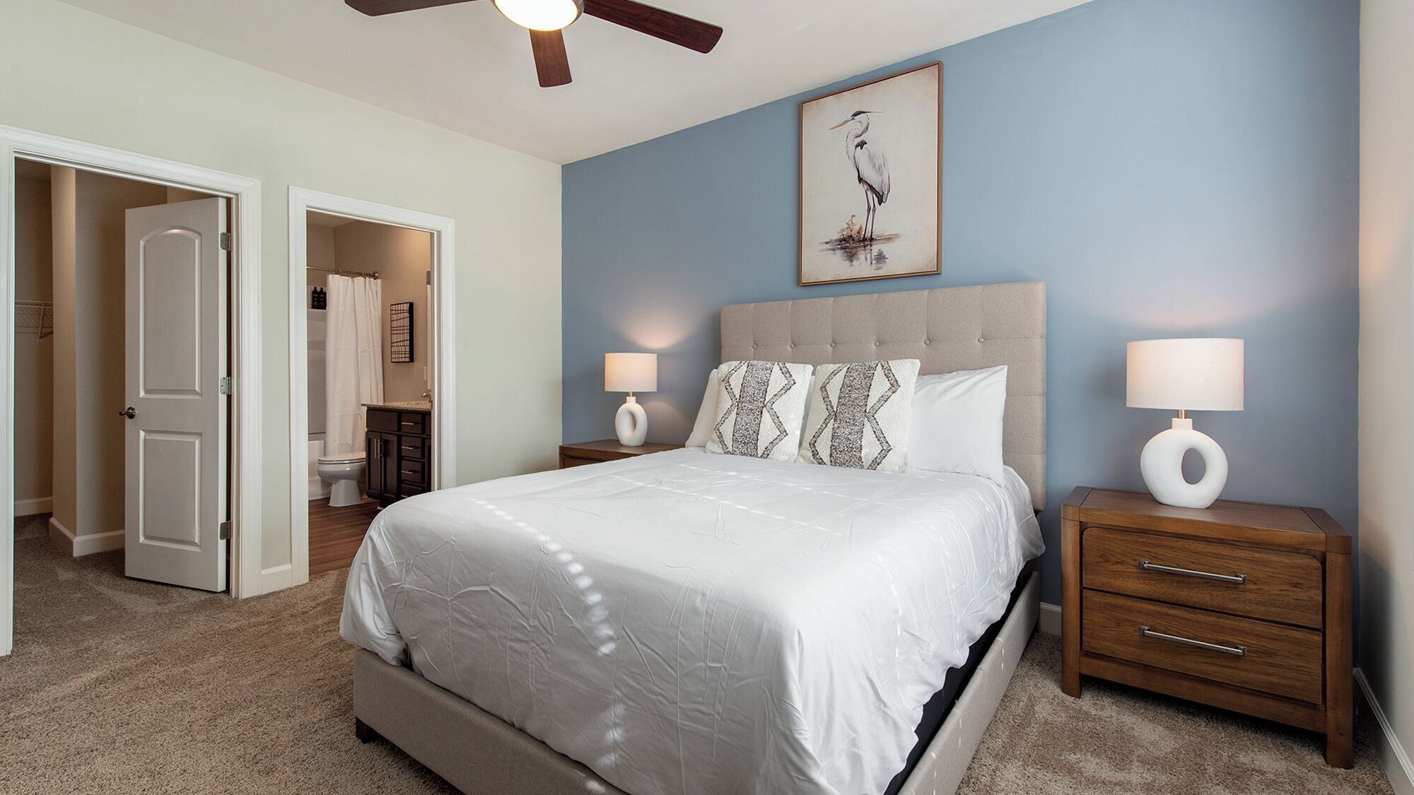 A bedroom with a bed, two bedside tables with lamps, a ceiling fan, and a framed artwork on the wall.