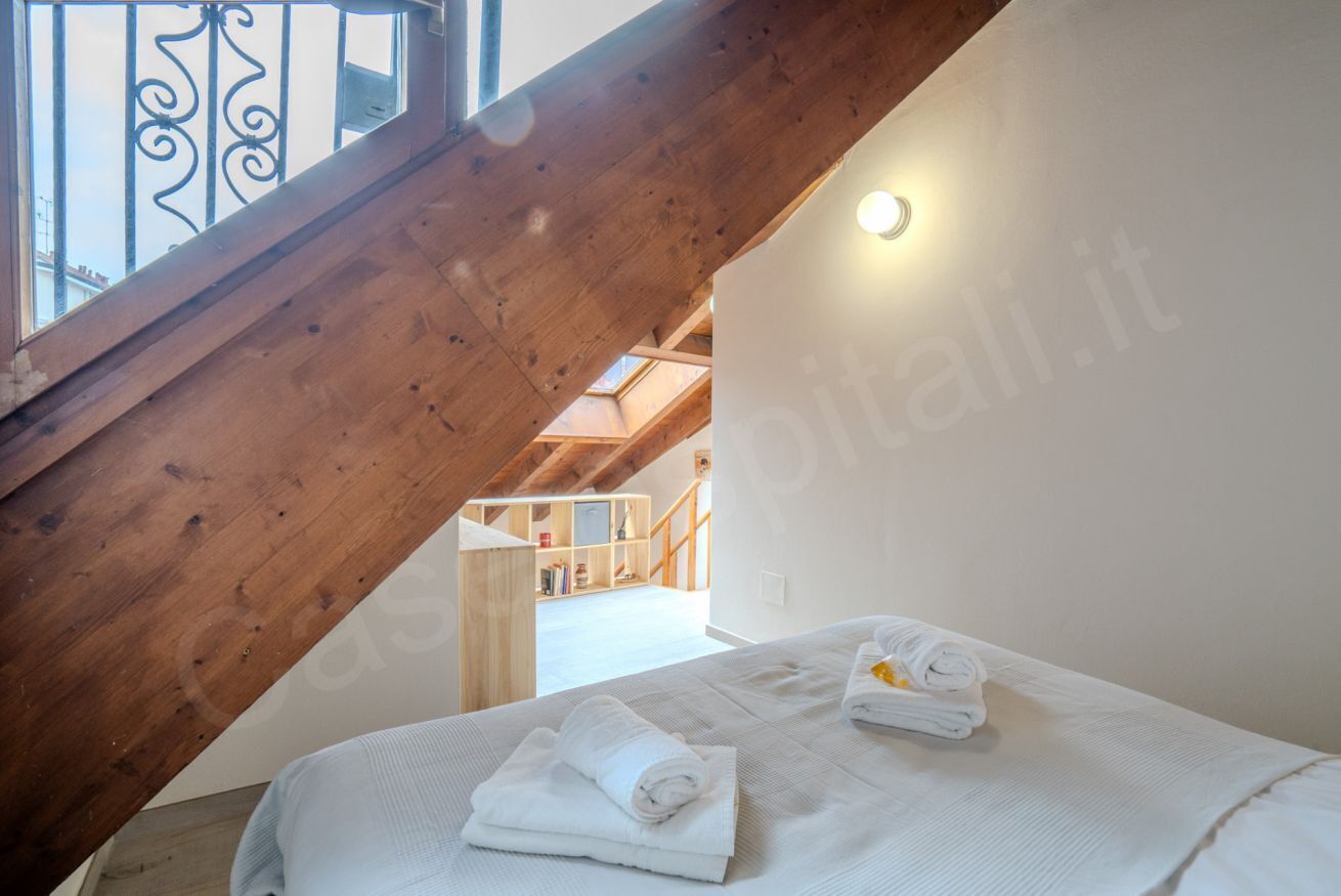 Une chambre avec un escalier en bois, un lit recouvert de draps blancs et une lucarne.