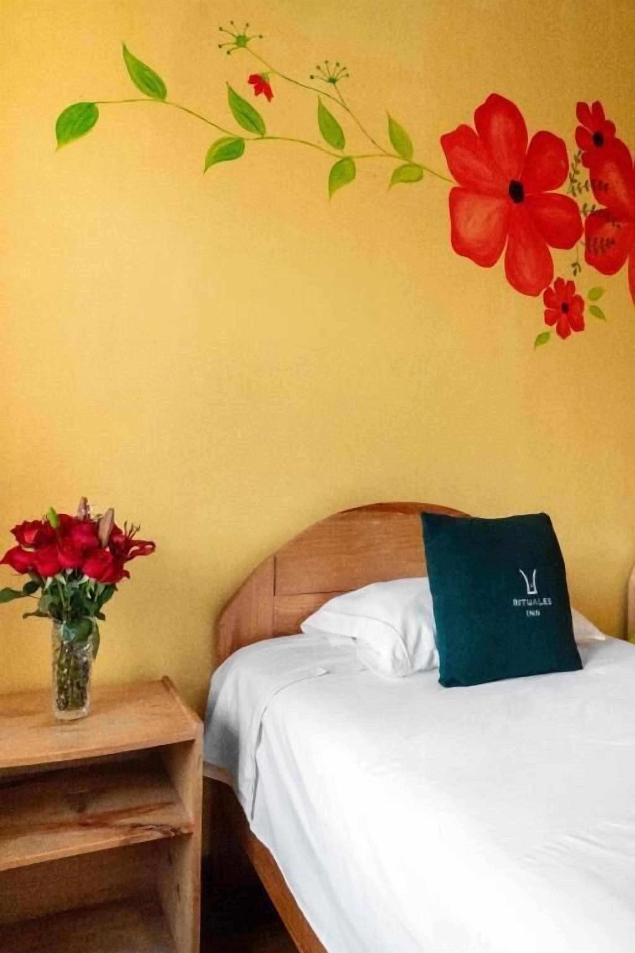 A hotel room with a bed, a nightstand with a vase of red flowers, and a wall mural of red flowers and green leaves.