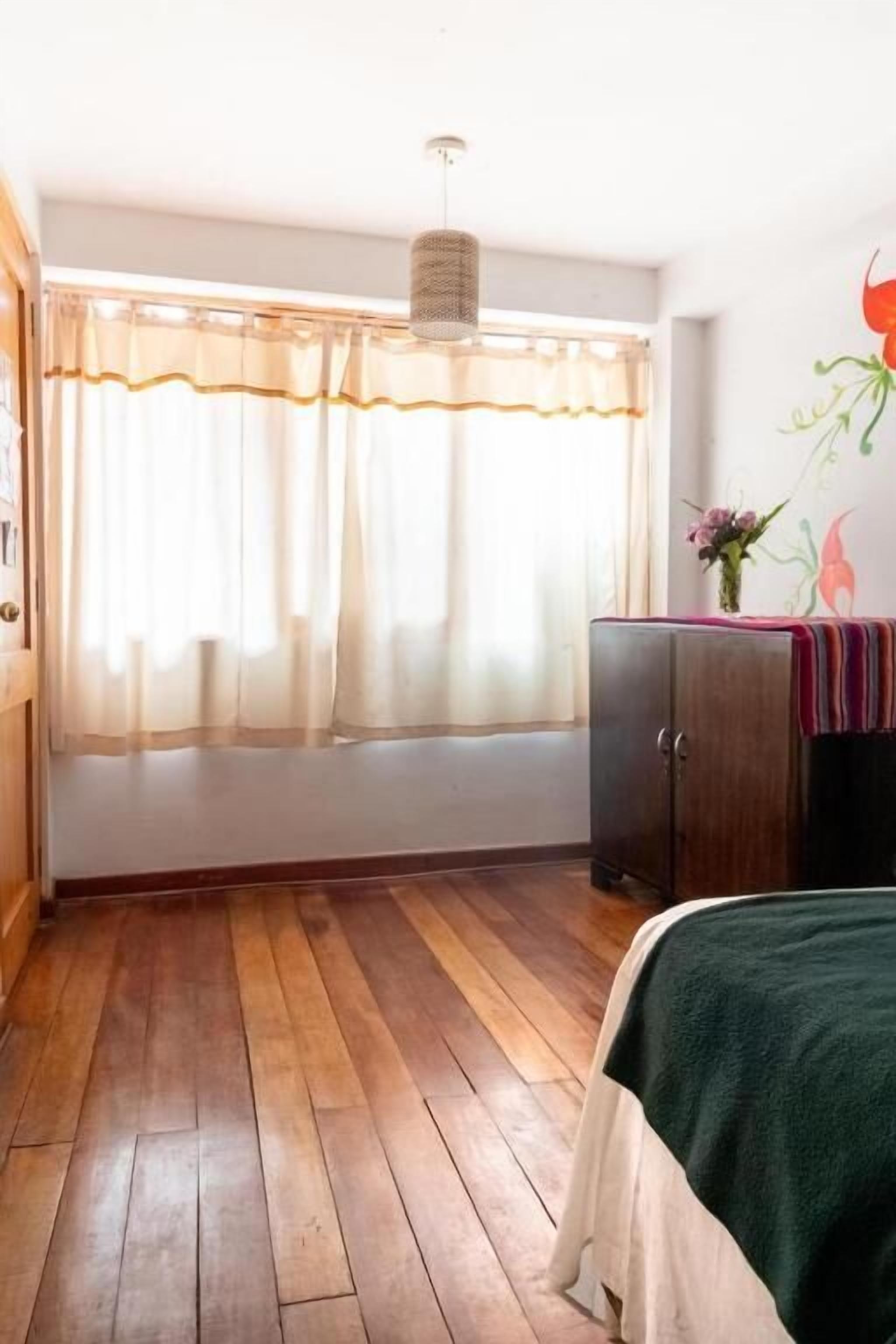 A room with a wooden floor, a bed with a green bedspread, a window with sheer curtains, and a hanging light fixture.