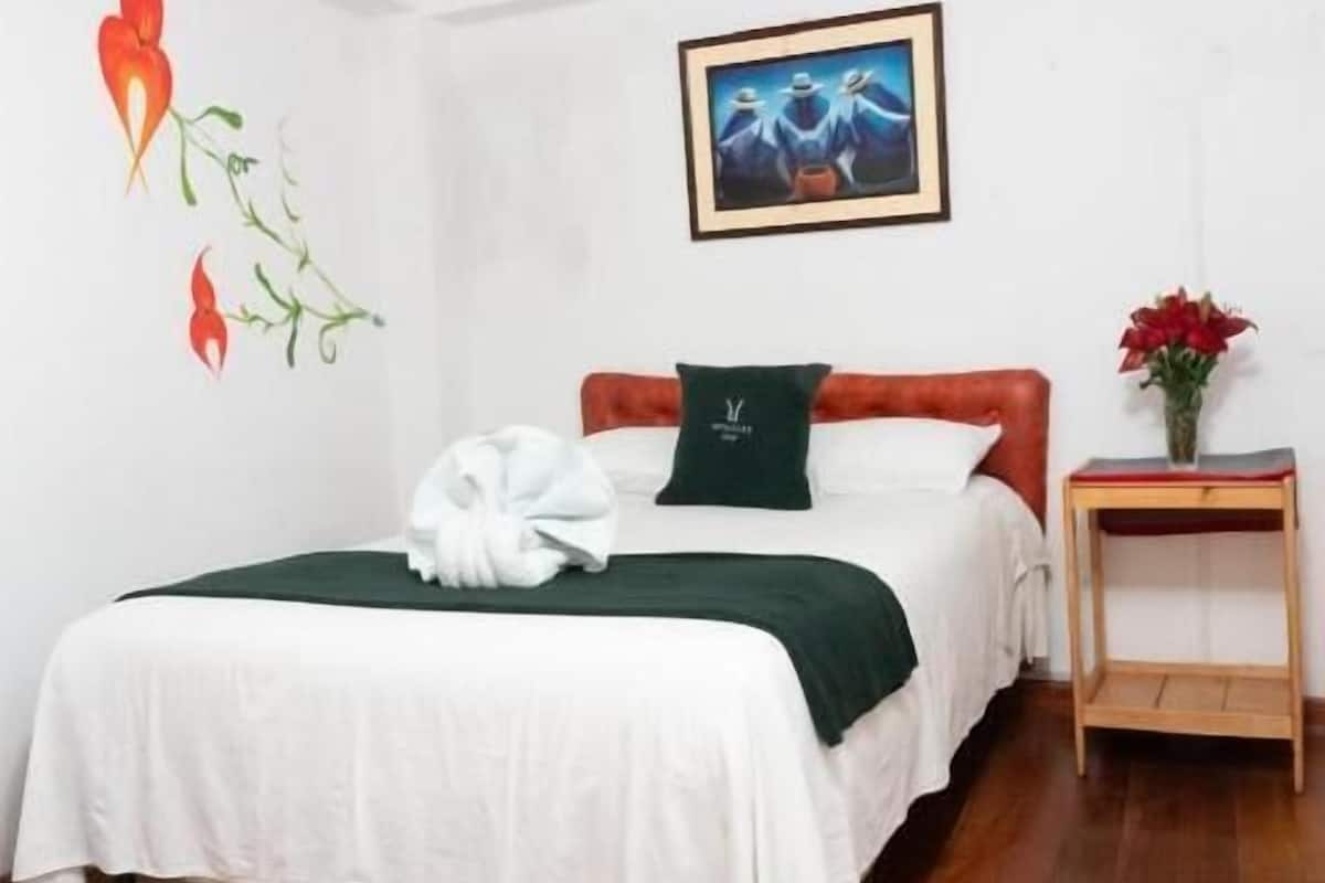 A neatly made bed with a green pillow and a white towel, a framed picture on the wall, and a small wooden nightstand with a vase of flowers.