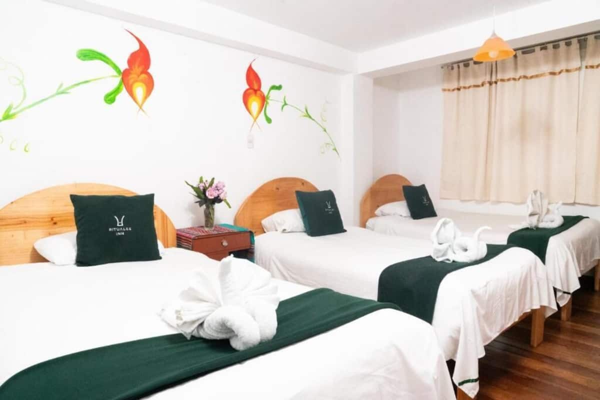 A hotel room with three beds, each with a green bedspread and matching pillows. There are wall art pieces and a vase with flowers on the nightstand.