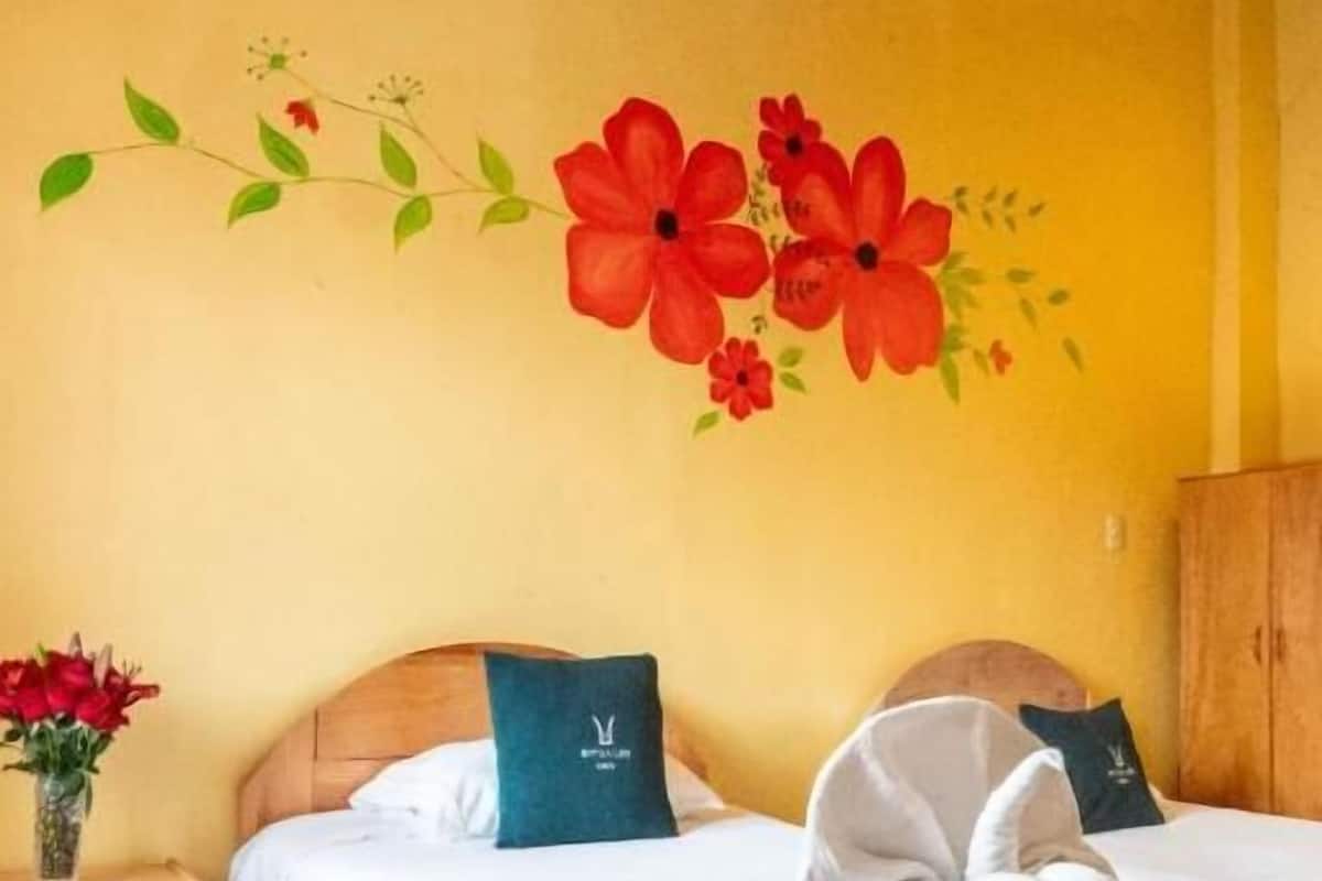 A hotel room with two beds, wooden headboards, a wooden wardrobe, a vase with red flowers, and a wall mural of red flowers.