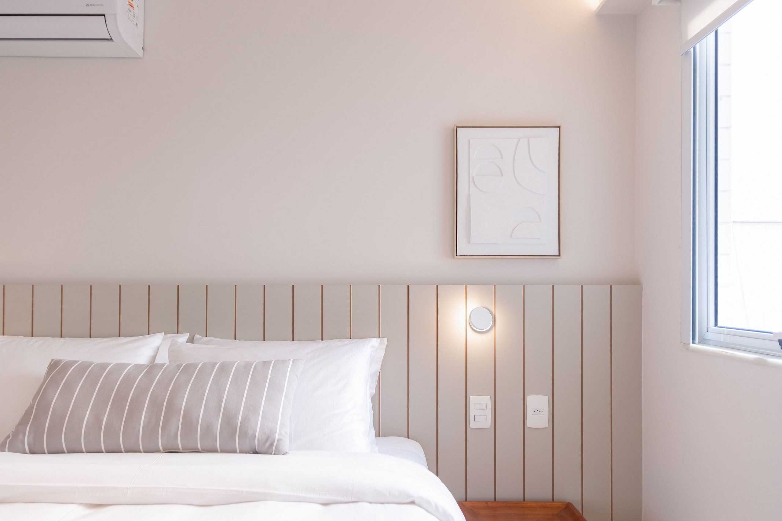 Una cama bien hecha con ropa de cama blanca y almohadas a rayas, un aire acondicionado de pared, una obra de arte abstracta enmarcada y una ventana con luz diurna.