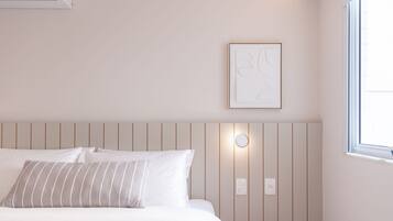 Una cama bien hecha con ropa de cama blanca y almohadas a rayas, un aire acondicionado de pared, una obra de arte abstracta enmarcada y una ventana con luz diurna.