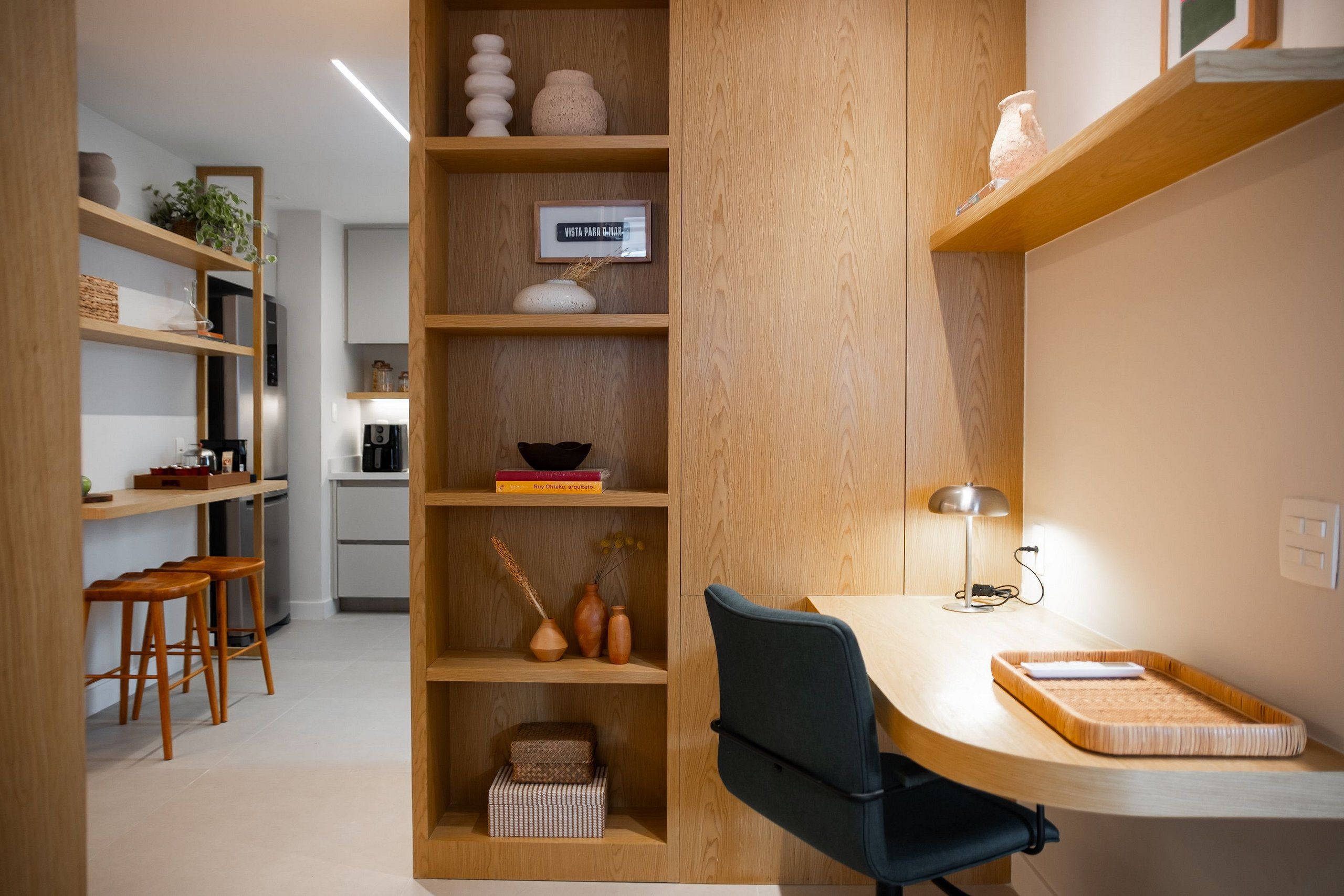 A modern kitchen with wooden shelves, a dining area with stools, and a workspace with a desk and lamp.
