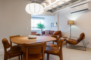 A modern dining area with a round wooden table, six chairs, a hanging light fixture, a potted plant, a sofa with cushions, and a floor lamp. - Leblon | 3 Quartos, 2 min da Praia - por Casapē Rio (Rio de Janeiro)