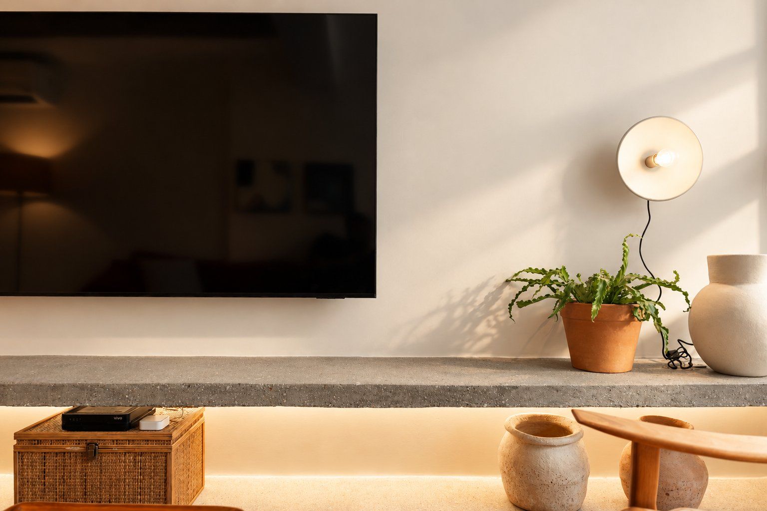 Een moderne woonkamer met een flatscreen televisie aan de muur, een kamerplant en een klein houten tafeltje met decoratieve objecten.