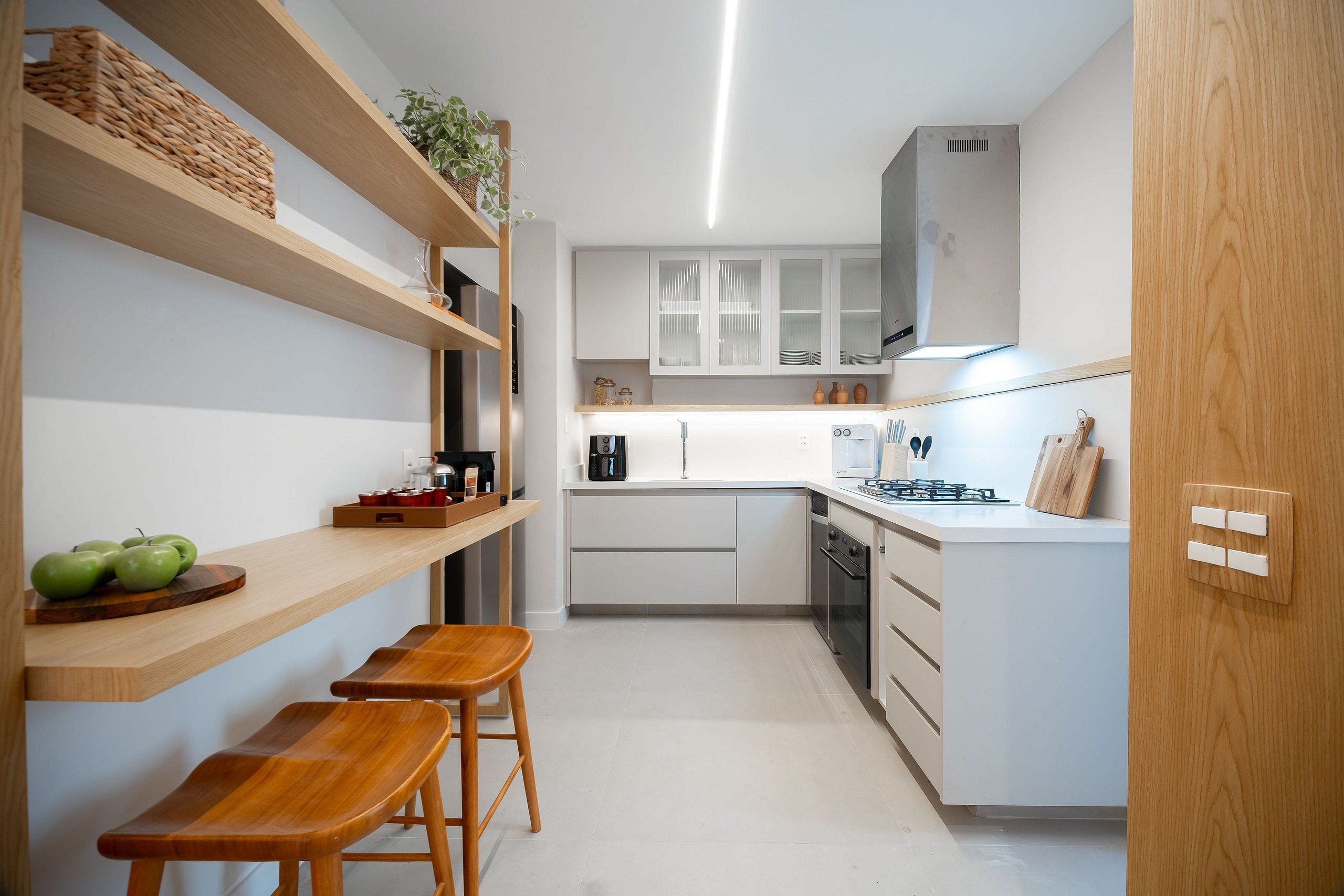 Una cocina compacta con armarios blancos, repisas de madera y un pequeño comedor con taburetes.