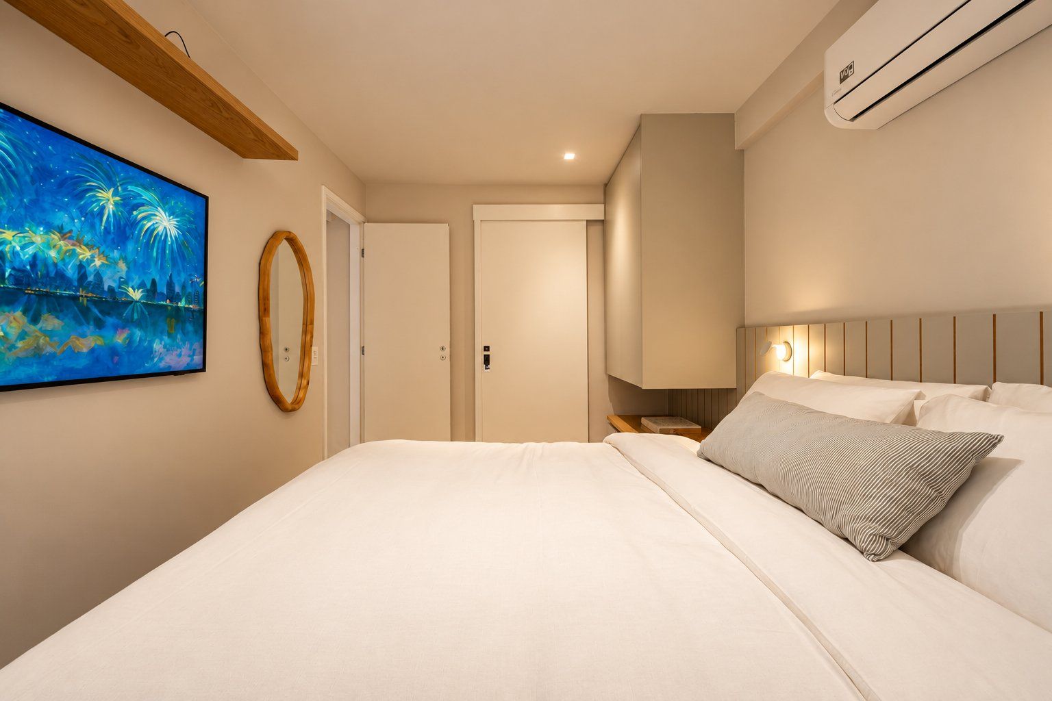 A hotel room with a large bed, a wall-mounted air conditioner, a painting, and a wooden ceiling beam.