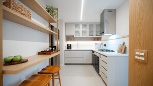 Μια μικρή κουζίνα με λευκά ντουλάπια, ξύλινα ράφια και μια μικρή τραπεζαρία με σκαμπό.