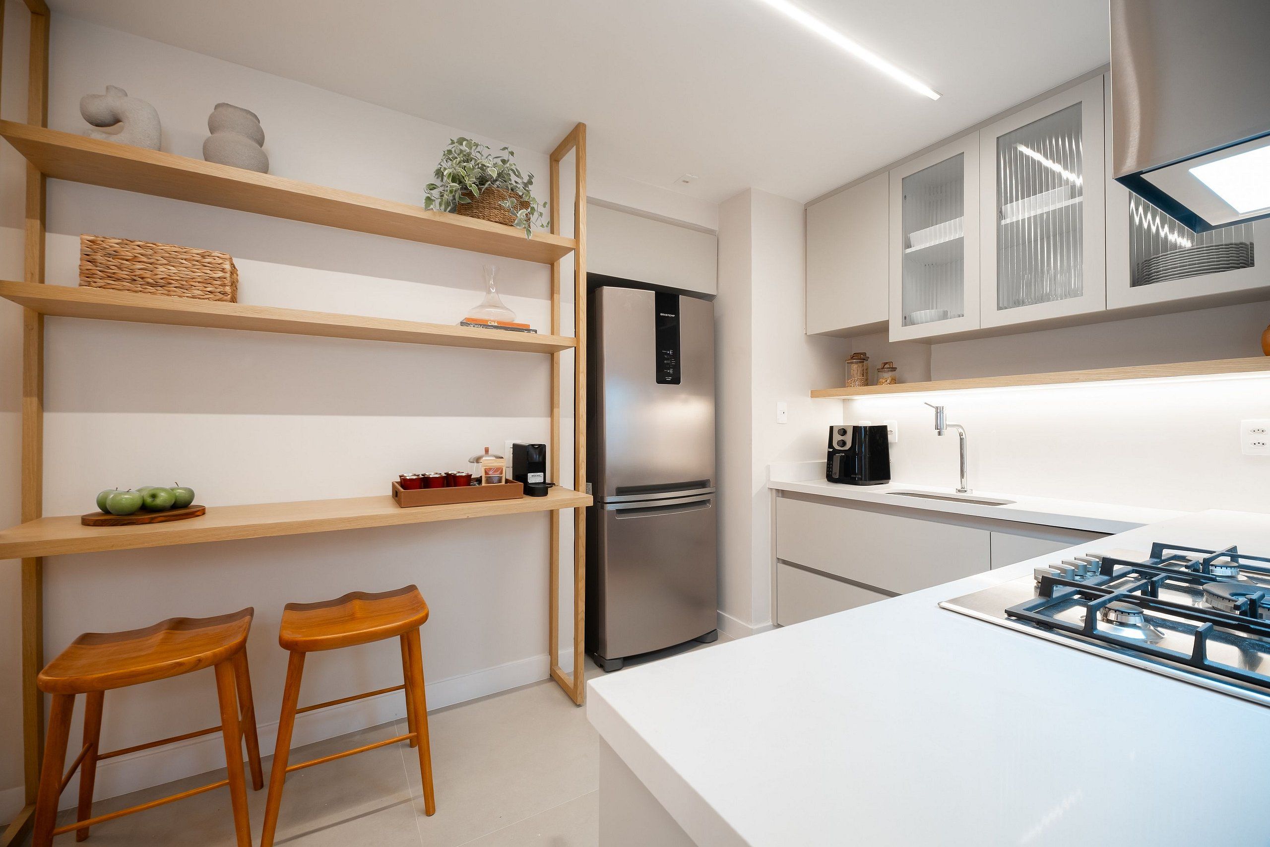 Una cocina moderna con refrigerador de acero inoxidable, estantes de madera, dos taburetes de madera y encimera blanca.