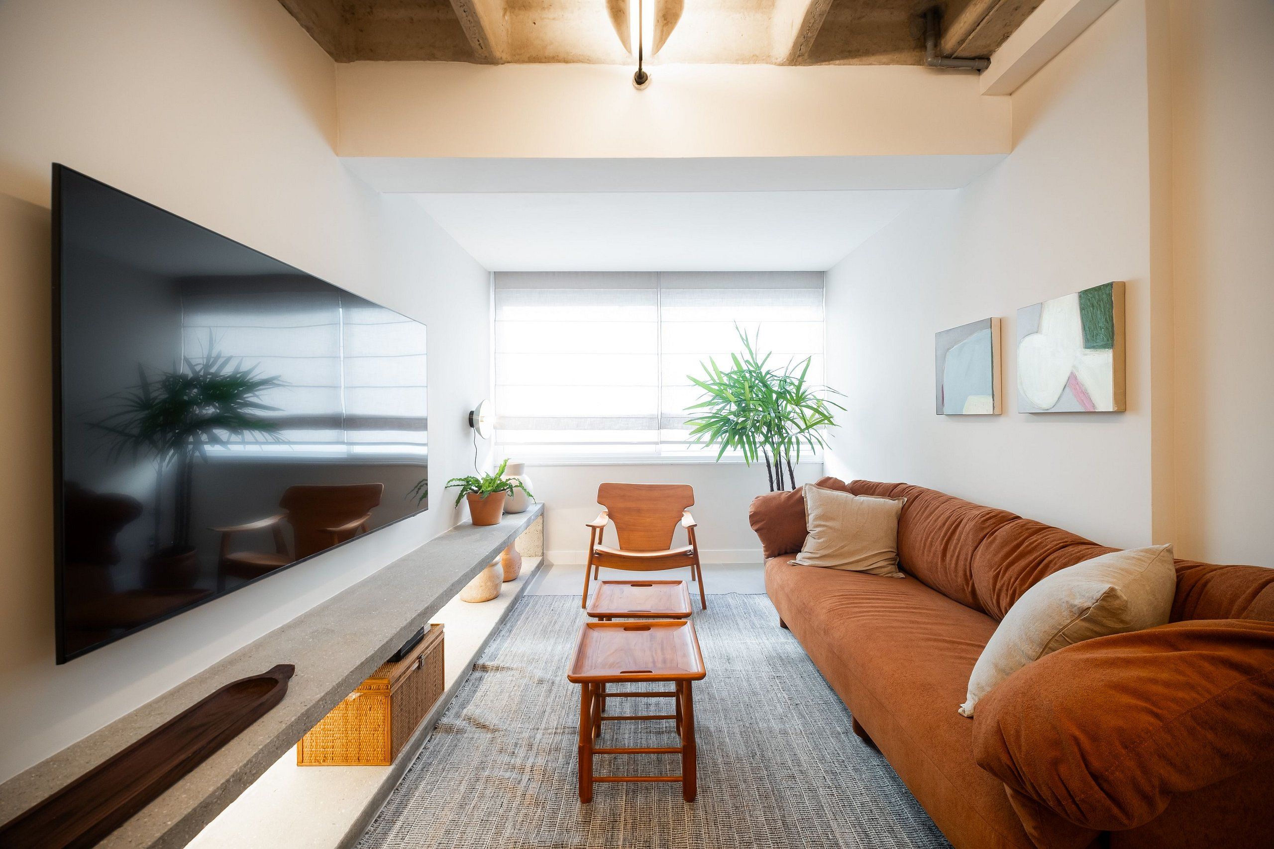A modern living room with a flat-screen TV, a wooden coffee table, a brown sofa, and a large window providing natural light.