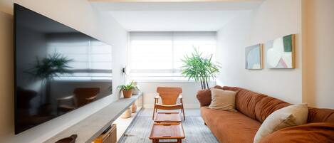 A modern living room with a flat-screen TV, a wooden coffee table, a brown sofa, and a large window providing natural light.