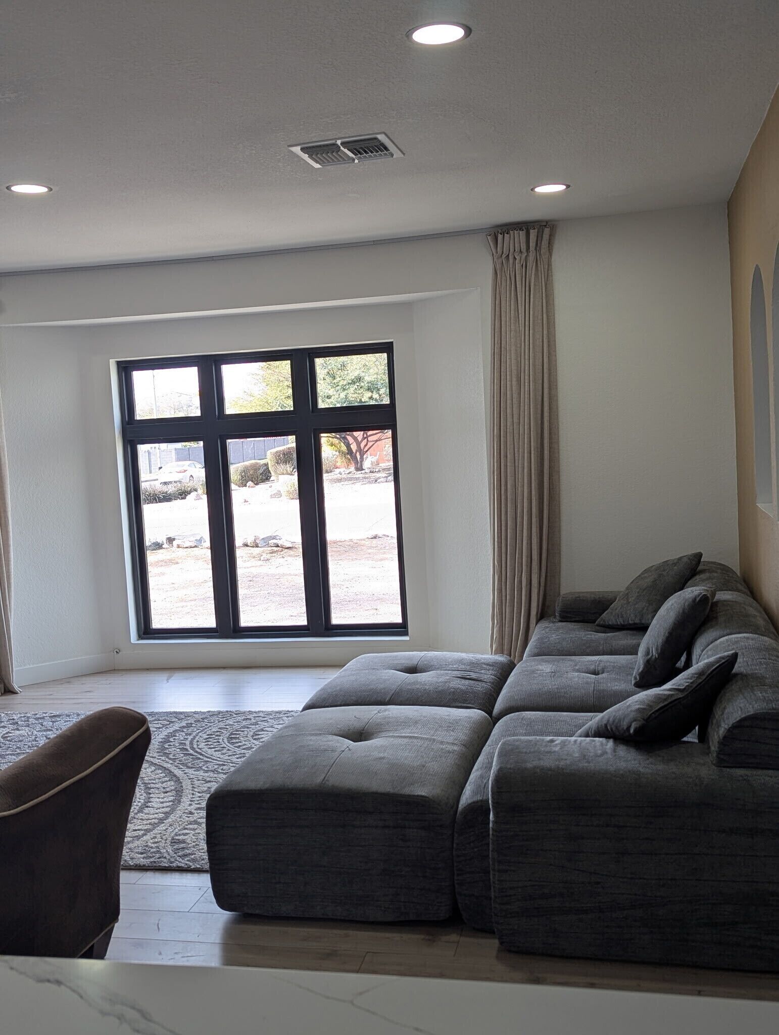 A spacious living room with a large sectional sofa, a small armchair, and a large window with a view of a landscaped area.