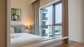 Et hotelværelse med en seng, et natbord, en lampe, et maleri og et stort vindue med udsigt til bygninger.