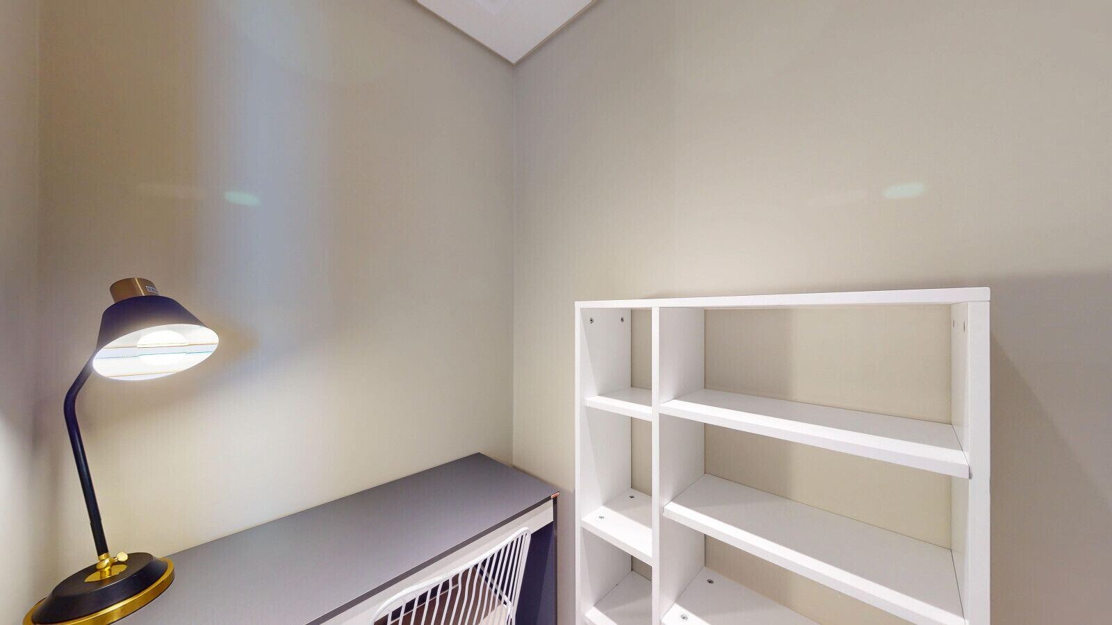 Um quarto com uma escrivaninha, uma luminária e uma estante branca na parede.